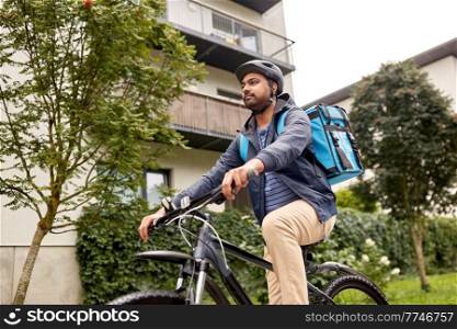 food shipping, profession and people concept - happy smiling indian delivery man in bike helmet with thermal insulated bag riding bicycle on city street. indian delivery man with bag riding bicycle
