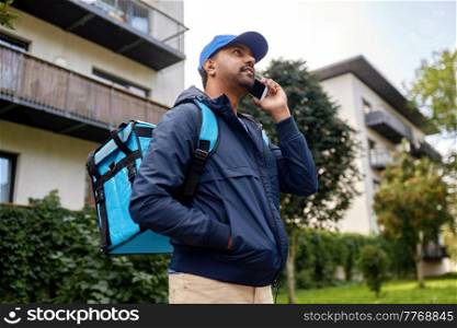 food shipping, profession and people concept - happy smiling indian delivery man with thermal insulated bag walking along city street and calling on smartphone. indian delivery man with bag calling on smartphone