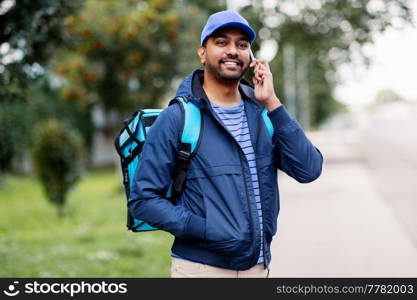 food shipping, profession and people concept - happy smiling indian delivery man with thermal insulated bag on city street calling on smartphone. indian delivery man with bag calling on smartphone