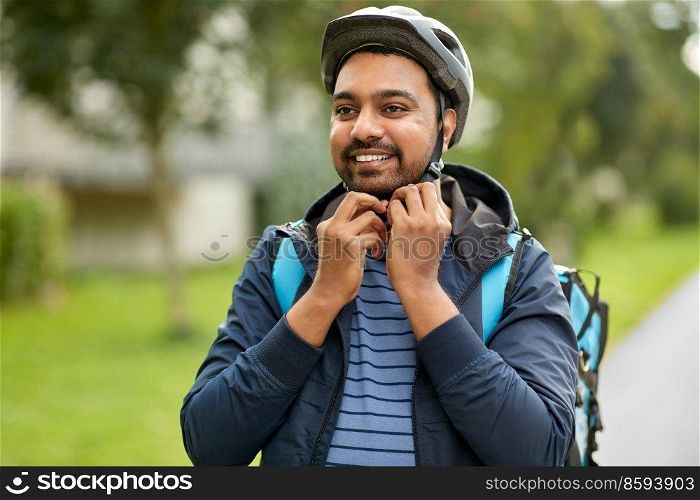 food shipping, safety and people concept - happy smiling delivery man with thermal insulated bag fastening his bicycle helmet on city street. food delivery man fastening bicycle helmet in city