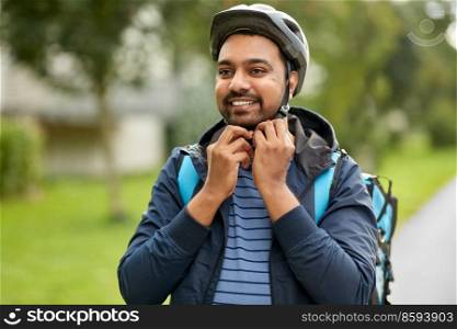 food shipping, safety and people concept - happy smiling delivery man with thermal insulated bag fastening his bicycle helmet on city street. food delivery man fastening bicycle helmet in city