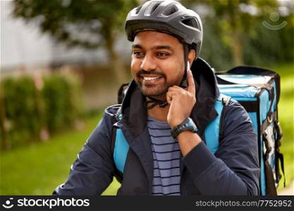 food shipping, technology and people concept - happy smiling delivery man in bicycle helmet with thermal insulated bag and earphones listening to music on city street. food delivery man in bicycle helmet with earphones