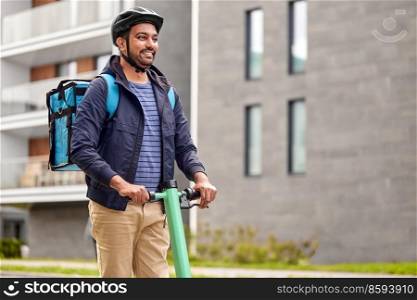 food shipping, transportation and people concept - happy smiling delivery man in bike helmet with thermal insulated bag riding electric scooter on city street. food delivery man with bag riding scooter