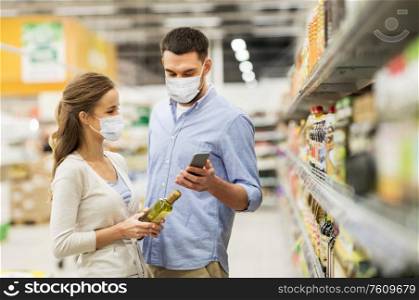 food shopping, consumerism and pandemic concept - happy couple wearing face protective medical masks for protection from virus disease with smartphone buying olive oil at grocery store or supermarket. couple in masks with phone and olive oil at store