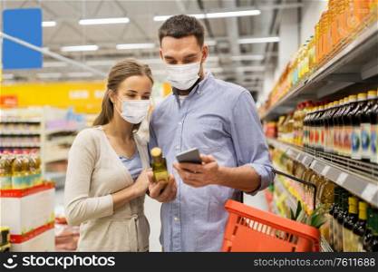 food shopping, consumerism and pandemic concept - happy couple wearing face protective medical masks for protection from virus disease with smartphone buying olive oil at grocery store or supermarket. couple in masks with phone and olive oil at store