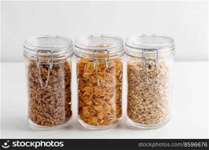 food storage, healthy eating and diet concept - jars with oat, corn flakes and granola on white background. close up of jars with oat, corn flakes and granola