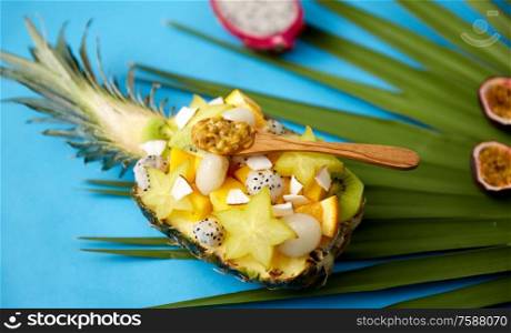 food, summer and healthy eating concept - mix of different exotic fruits in pineapple with wooden spoon on green fan palm leaf and blue background. mix of exotic fruits in pineapple with spoon