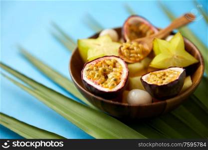 food, summer and healthy eating concept - mix of different exotic fruits in wooden plate with spoon and green fan palm leaf on blue background. mix of exotic fruits in wooden plate with spoon