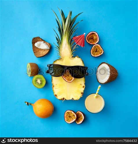 food, summer and healthy eating concept - pineapple in black sunglasses, drink with paper straw and other exotic fruits on blue background. pineapple in sunglasses with other exotic fruits