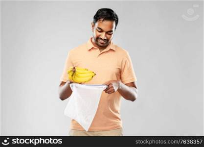 food, sustainability and eco living concept - happy smiling young indian man in polo t-shirt putting bananas into reusable string bag over grey background. happy man putting bananas into reusable string bag
