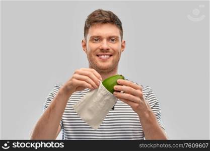 food, sustainability and eco living concept - smiling young man in striped t-shirt with avocado in reusable canvas bag over grey background. smiling man with avocado in reusable canvas bag