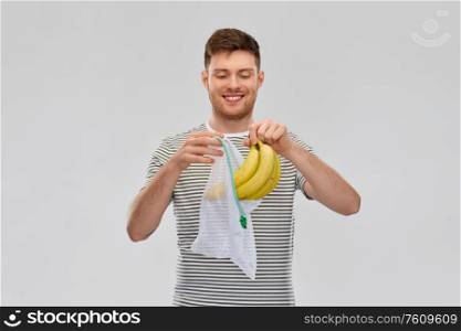 food, sustainability and eco living concept - smiling young man in striped t-shirt putting bananas into reusable string bag over grey background. smiling man putting bananas into reusable net bag