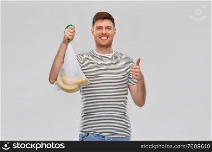 food, sustainability and eco living concept - smiling young man in striped t-shirt holding reusable string bag with bananas and showing thumbs up over grey background. happy man holding reusable string bag with bananas