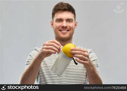 food, sustainability and eco living concept - smiling young man in striped t-shirt with lemon in reusable canvas bag over grey background. smiling man with lemon in reusable canvas bag