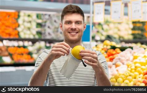 food, sustainability and eco living concept - smiling young man in striped t-shirt with lemon in reusable canvas bag over grocery store or supermarket background. smiling man putting lemon in reusable grocery tote