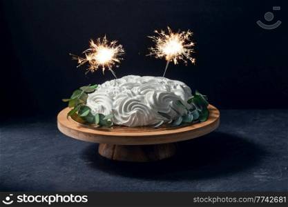 food, sweets and objects concept - close up of meringue or zephyr cake with sparklers on wooden stand with eucalyptus branch over dark background. close up of zephyr cake with sparklers on stand