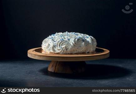 food, sweets and objects concept - meringue or zephyr cake with blue sprinkles on wooden stand over dark background. close up of zephyr cake on wooden stand