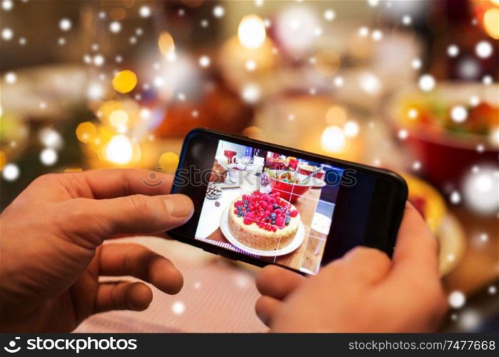 food, technology and holidays concept - close up of male hands photographing cake by smartphone at christmas dinner over snow. hands photographing food at christmas dinner
