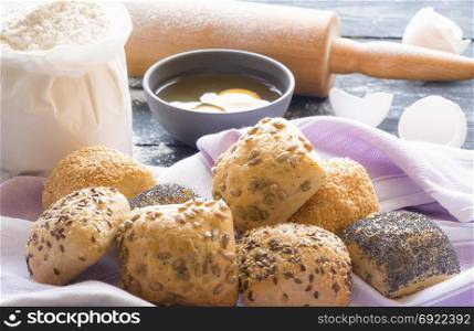 Food theme image with homemade bread rolls with sesame, poppy seeds, sunflower and flax seeds on a towel and the ingredients for their baking in the background.