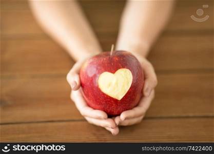 food, valentines day and health concept - close up of hands holding ripe red apple with carved heart shape over wooden table. close up of hands holding apple with carved heart