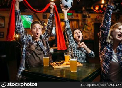 Football fans with scarf and ball in hands watching match and drinks beer in sports bar. Tv broadcasting, young friends leisures in pub, favorite team wins. Football fans with scarf and ball in sports bar