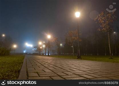 Footpath in city park at night in fog with streetlights. Beautiful foggy evening in the autumn alley with burning lanterns.. Footpath in city park at night in fog with streetlights. Beautiful foggy evening in the autumn alley with burning lanterns