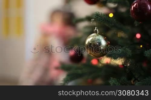 Foreground glass baubles on Christmas tree&acute;s branch and blurry happy mother and daughter embracing, having fun and playing patty cake while enjoying winter holidays at home. Dolly shot.