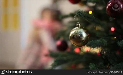Foreground glass baubles on Christmas tree&acute;s branch and blurry happy mother and daughter embracing, having fun and playing patty cake while enjoying winter holidays at home. Dolly shot.