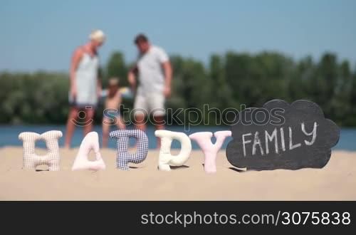 Foreground word happy made of fabric padded letters and word family written with chalk on a black board. Blurred background of family with kid playing on the beach during summer vacation