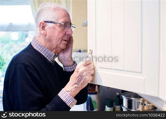 Forgetful Senior Man With Dementia Looking In Cupboard At Home