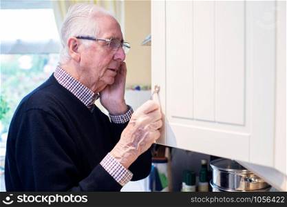 Forgetful Senior Man With Dementia Looking In Cupboard At Home