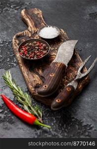 Fork and knife on chopping board with pepper and salt with rosemary on black kitchen board.