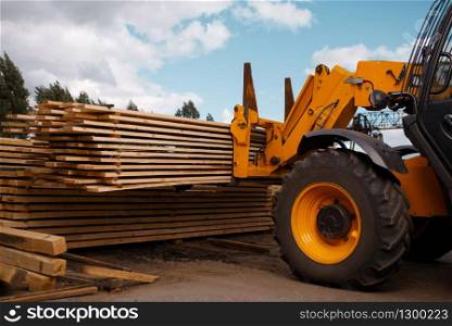 Forklift loads the boards in the lumber yard outdoor. Autoloader works on timber mill warehouse, woodworking industry, carpentry. Wood processing on factory, forest sawing, sawmill. Forklift loads the boards in the lumber yard