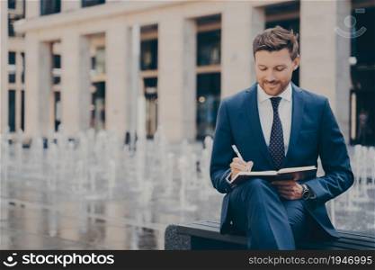 Formally dressed male office worker organising his day in notebook, writing it all down while sitting on bench, fountains working behind him, handsome businessman enjoying work outdoors on weekend. Formally dressed office worker organising his day in note book while working outdoors