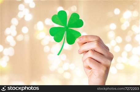 fortune, luck and st patricks day concept - hand holding green paper shamrock over festive lights on beige background. hand holding green paper four-leaf clover