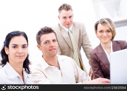 Four business colleagues working together on laptop computer in office, looking at camera, smiling.