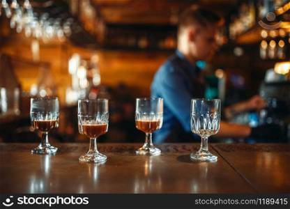 Four glasses standing on bar counter, male bartender in apron on background. Barman occupation, bartending. Glasses on bar counter, bartender on background
