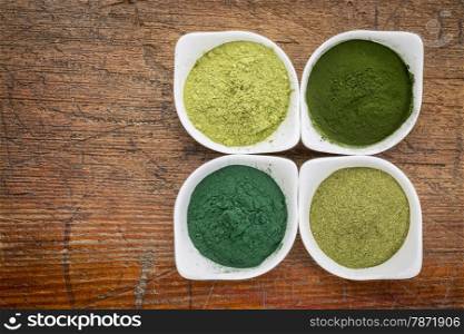 four healthy green dietary supplement powders (spirulina, chlorella, wheatgrass and moringa leaf) in white bowls on a grunge wood