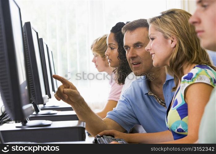 Four students sitting at computer terminals with teacher helping one of them (depth of field/high key)