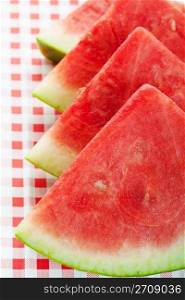 Four watermelon wedges on a red, checkered picnic table cloth. Shallow depth of field.