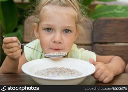 Four-year-girl at the table in the fresh air blowing on a spoonful of porridge