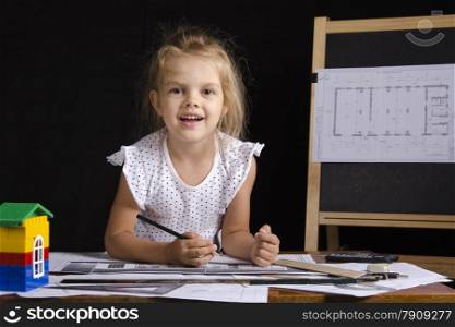 Four-year-old girl in the image of the architect. The girl sitting at the table with the drawings. In the background stands the Board with the drawing. Girl fun looking at the picture