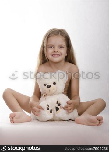 Four-year-old girl sitting planted bear in front of him and embraces him. Studio light background