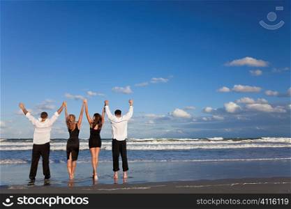 Four young people, two couples, holding hands, arms raised having fun and celebrating on a beach