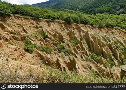 Fragment of the famous Stob Pyramids with unusual shape red and yellow rock formations, green bushes and trees around, west share of Rila mountain, Kyustendil region, Bulgaria, Europe