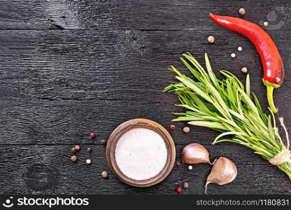 Frame of pink Himalayan salt in bowl, peas of colored pepper, a bunch of fresh rosemary, a pod of red hot pepper and two cloves of garlic on black wooden board on top