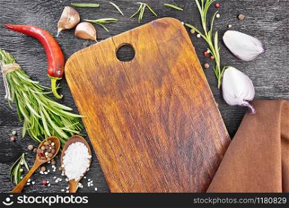 Frame of wooden dill, spoons with peas of pepper and salt, sprigs of fresh rosemary, pods of red hot pepper, slices of garlic, onions and a napkin on black wooden board top