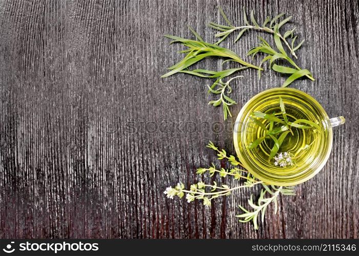 Frame with herbal tea in a glass cup, sprigs of tarragon, rosemary and thyme on the background of dark wooden board on top