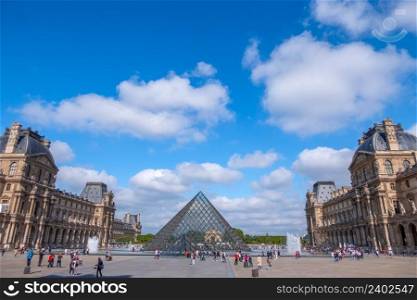France. Sunny summer weather in Paris. The famous courtyard of the Louvre Museum and the glass pyramids. Many tourists. Clouds. Louvre Courtyard With Tourists and Light Clouds in the Blue Sky