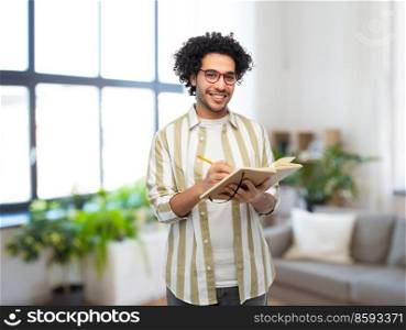 freedom, motion and happiness concept - happy smiling young man in glasses with diary and pencil writing over home room background. happy smiling man with diary and pencil at home